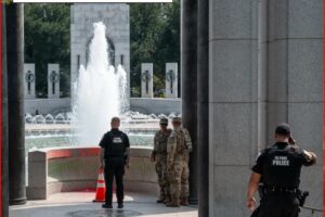 Uniformed police officers stand near a fountain at a memorial, with tall stone pillars surrounding.