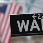 Close-up of a Wall Street street sign with blurred U.S. flags in the background.