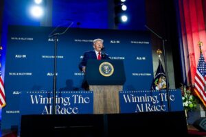 Donald Trump speaks behind a podium at the White House AI Summit, flanked by “Winning the AI Race” signs.