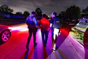 ICE officer and FBI agent escort a handcuffed man during a nighttime operation in Dallas.