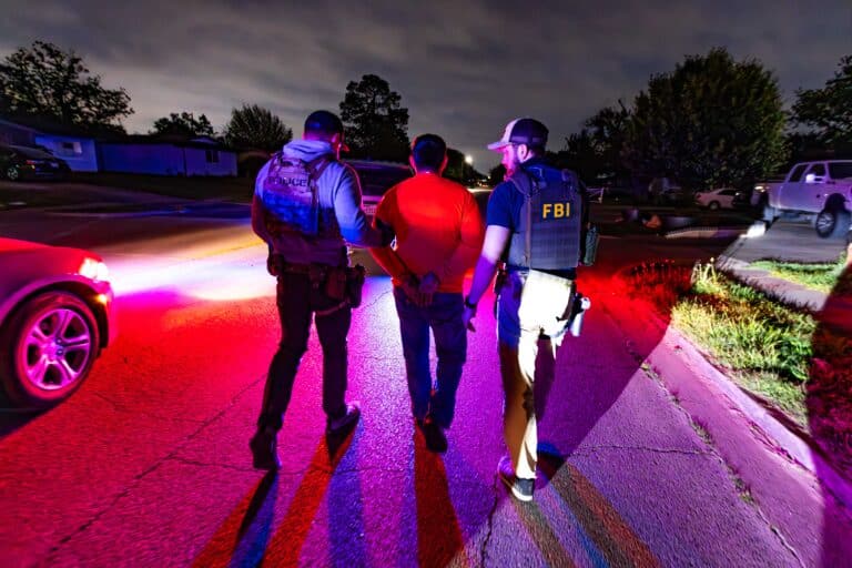 ICE officer and FBI agent escort a handcuffed man during a nighttime operation in Dallas.
