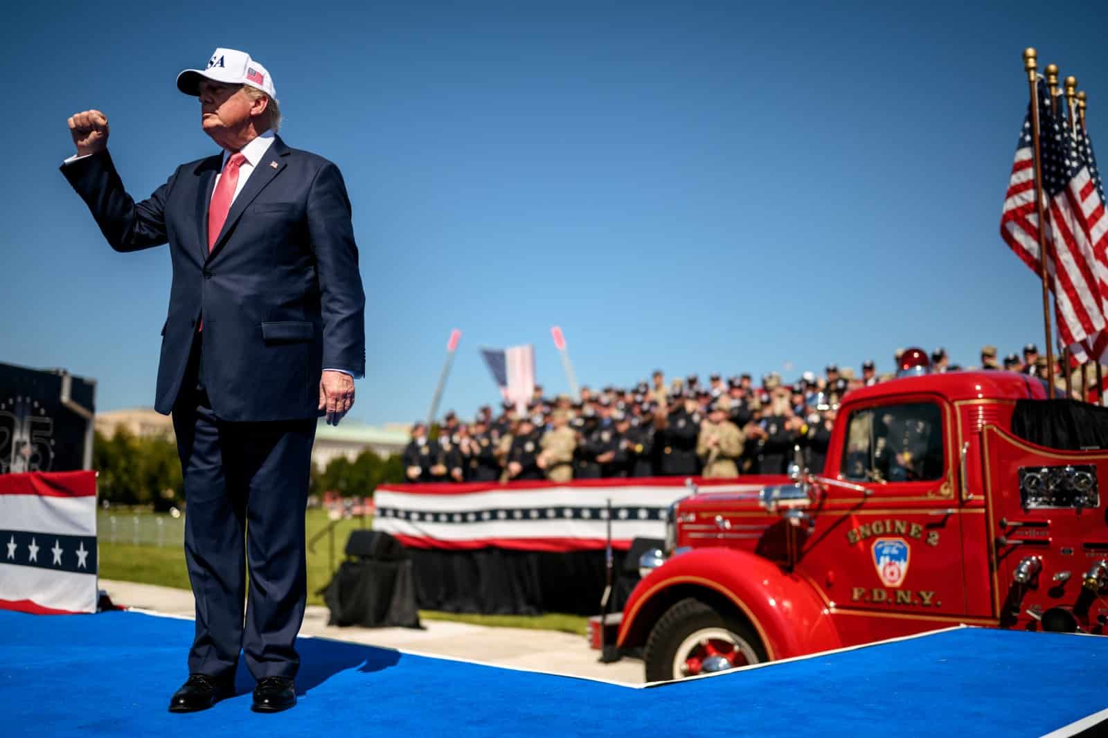 Donald Trump raises his fist on an outdoor stage with American flags behind him.