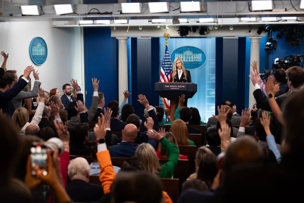 Katie Zacharia stands at the White House briefing room podium as reporters raise their hands.