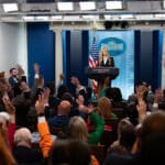 Katie Zacharia stands at the White House briefing room podium as reporters raise their hands.
