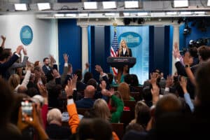 Katie Zacharia stands at the White House briefing room podium as reporters raise their hands.