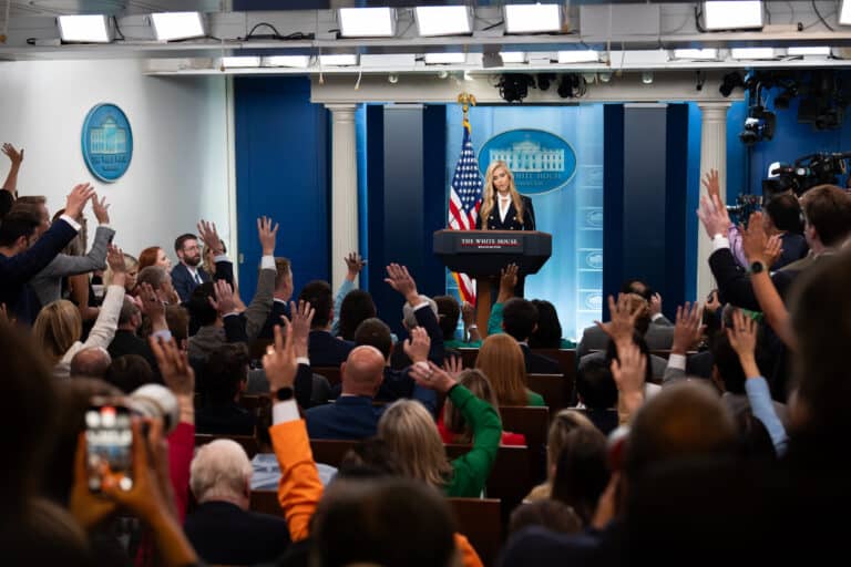 Katie Zacharia stands at the White House briefing room podium as reporters raise their hands.