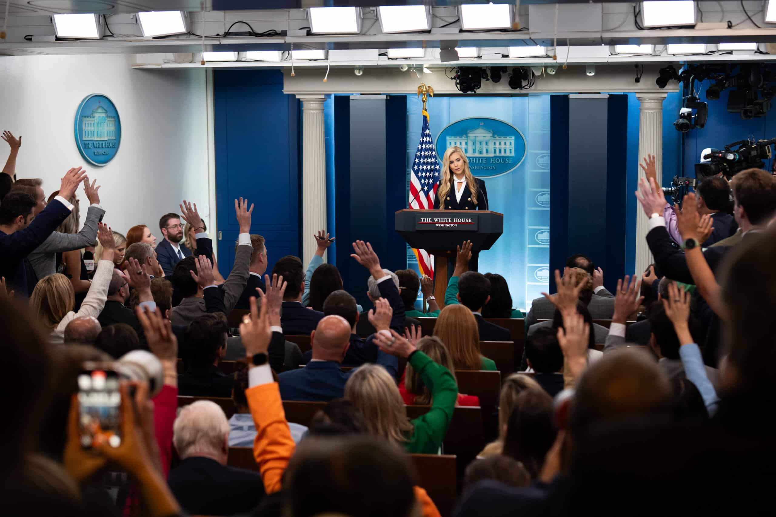 Katie Zacharia stands at the White House briefing room podium as reporters raise their hands.