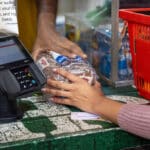 Shopper places a loaf of bread beside a payment terminal at a grocery checkout.