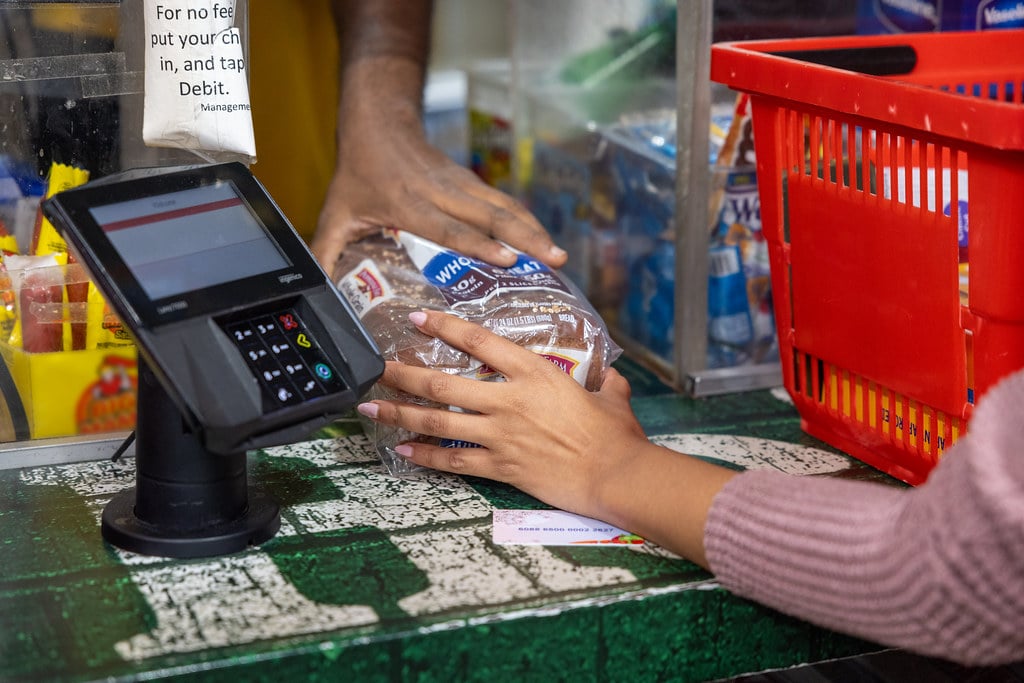 Shopper places a loaf of bread beside a payment terminal at a grocery checkout.
